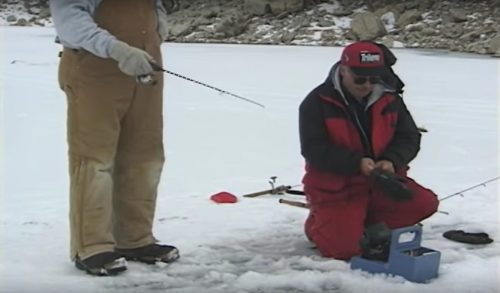 Two men ice fishing on a snowy lake, one kneeling and checking equipment, the other standing nearby.