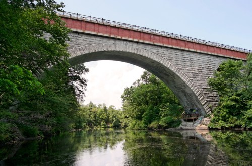 A large stone arch bridge spans over a calm river, surrounded by lush green trees under a clear blue sky.
