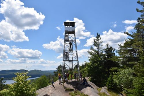 A tall fire tower on a rocky outcrop, surrounded by trees and people, under a bright blue sky with fluffy clouds.