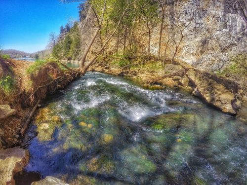 A serene river flows over rocks, surrounded by lush greenery and steep cliffs under a clear blue sky.
