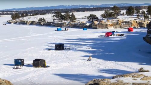 A snowy landscape with ice fishing tents scattered across a frozen lake, surrounded by trees and rocky cliffs.