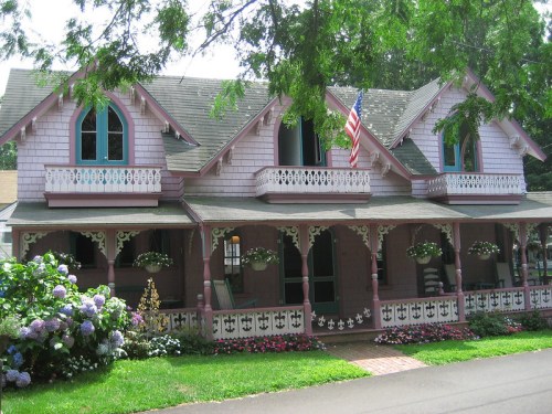A charming pink Victorian house with decorative trim, an American flag, and colorful flower beds in front.