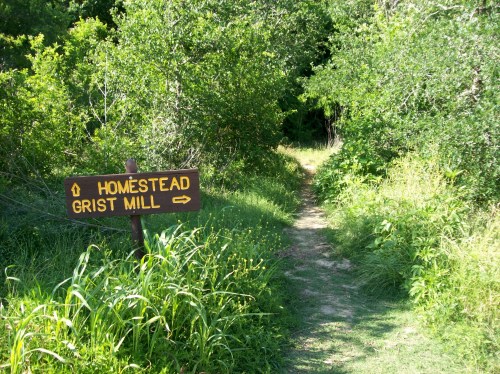 A wooden sign points to a path leading to the Homestead Grist Mill, surrounded by lush greenery.