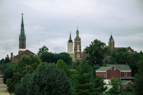 A scenic view of a church complex with multiple steeples, surrounded by trees and buildings under a cloudy sky.
