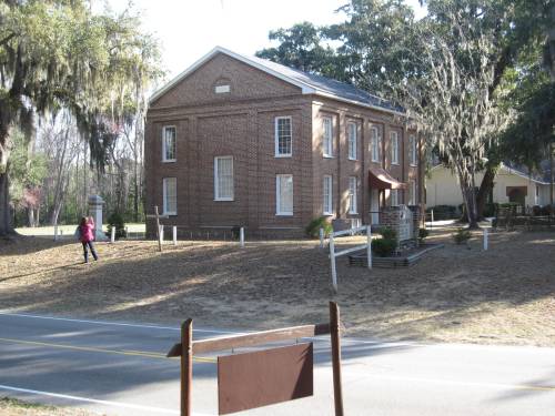 A brick building with windows and a sloped roof, surrounded by trees and a dirt path, with a person walking nearby.
