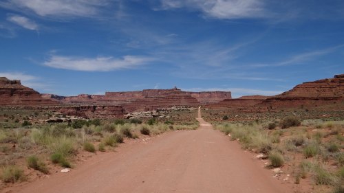 A dirt road leads through a desert landscape with red rock formations and a blue sky dotted with clouds.