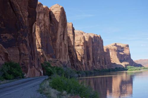 A scenic view of towering red rock cliffs beside a calm river, with a road running alongside the landscape.