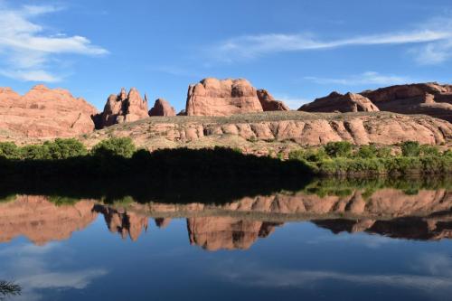 A serene landscape featuring red rock formations reflected in calm water under a blue sky with scattered clouds.
