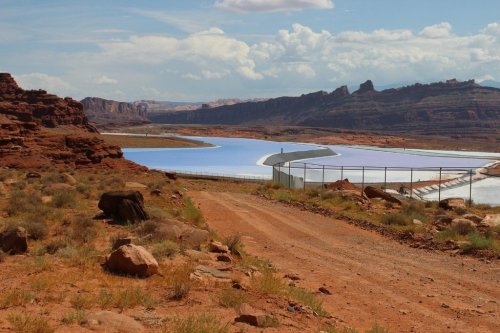 A desert landscape with a winding blue reservoir surrounded by rocky cliffs and a dirt path leading into the scene.