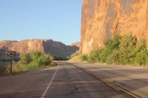 A winding road beside towering red rock formations and greenery, with a sign for "Indian Writing" visible.