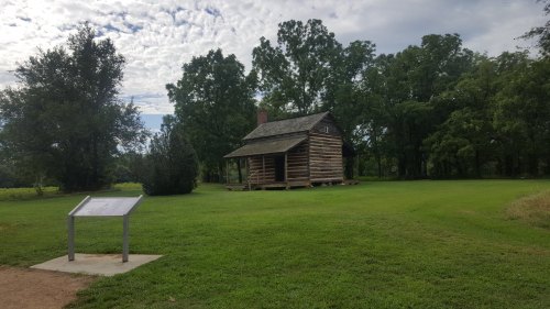 A rustic log cabin surrounded by green grass and trees, with an informational sign nearby under a cloudy sky.