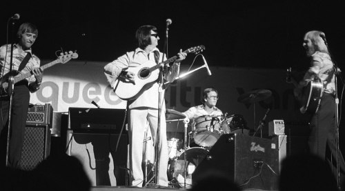 A black and white photo of a band performing on stage with guitars and drums, featuring four musicians in stylish outfits.