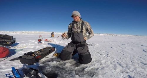 A person in winter gear kneels on ice, fishing through a hole, with equipment and another person in the background.