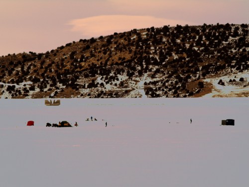 A snowy landscape with small tents and people ice fishing on a frozen lake, surrounded by hills and a pastel sky.