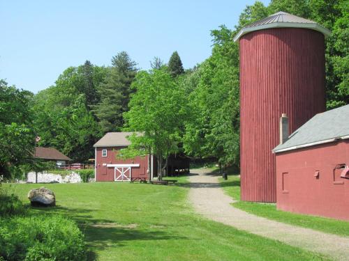 A scenic farm with red barns and a silo, surrounded by green trees and a gravel path on a sunny day.