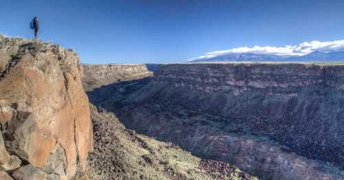 A person stands on a rocky cliff overlooking a vast canyon under a clear blue sky.