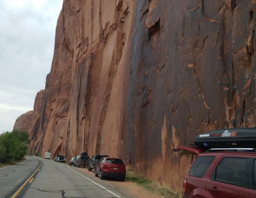 Cars parked along a road beside towering red rock formations under a cloudy sky.