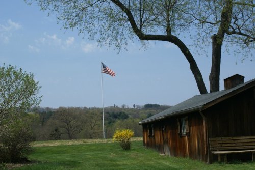 A wooden cabin with a bench, a large tree, and an American flag waving in the background against a clear sky.
