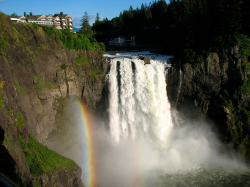 A majestic waterfall cascades down rocky cliffs, with a vibrant rainbow arcing through the mist. Lush greenery surrounds the scene.