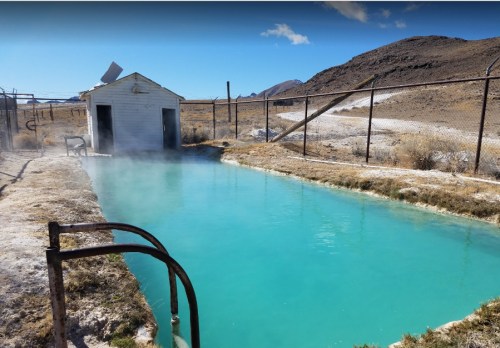 A steaming turquoise hot spring beside a small white building, surrounded by a barren landscape and mountains.