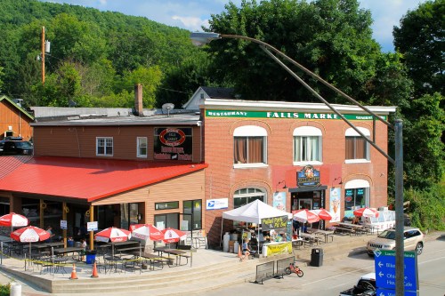A brick building with a red roof, featuring outdoor seating and umbrellas, surrounded by greenery and a streetlight.