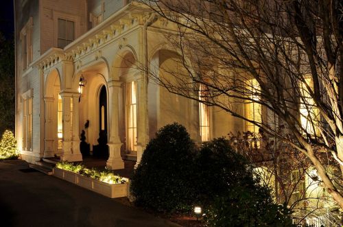 A beautifully lit historic building entrance at night, featuring arches, greenery, and warm glowing windows.