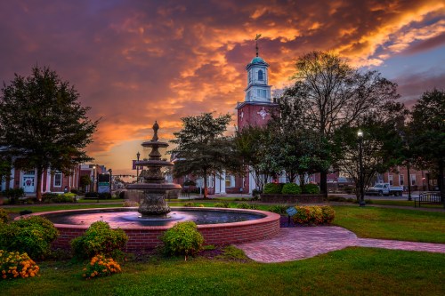 A scenic sunset over a park with a fountain and trees, featuring a historic building in the background.