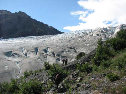 Two hikers traverse a rocky landscape near a large glacier under a clear blue sky.