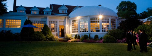 A beautifully lit house with large windows, surrounded by greenery, and people mingling in the garden at dusk.
