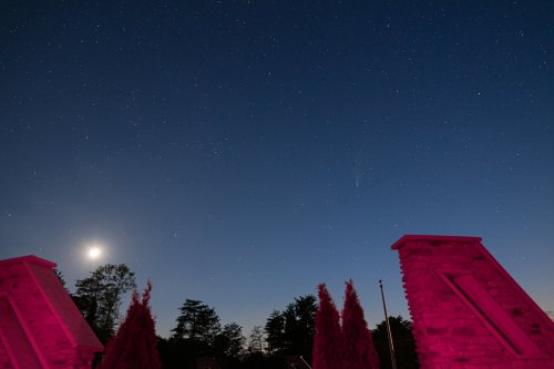 A night sky filled with stars, featuring a bright moon and a comet, framed by stone structures and trees.