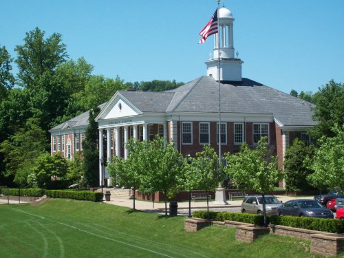 A brick building with a white dome and flag, surrounded by green trees and a lawn, on a sunny day.