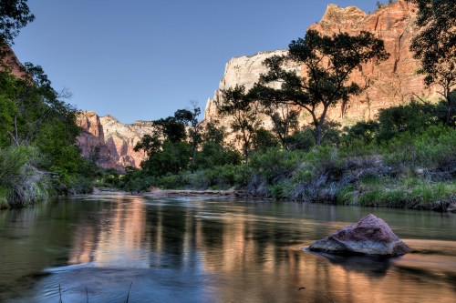 A serene river flows through a canyon, reflecting cliffs and trees under a clear blue sky.