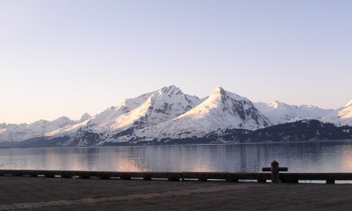 Snow-capped mountains reflect in calm water during sunrise, with a clear sky above.