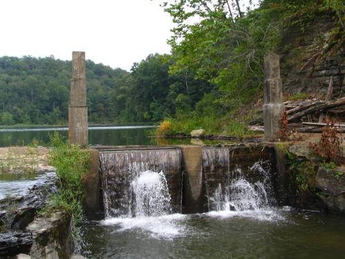 A serene landscape featuring a small waterfall flowing over stone structures, surrounded by lush greenery and calm water.