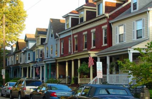 A row of colorful houses with porches and American flags, lined along a street with parked cars.