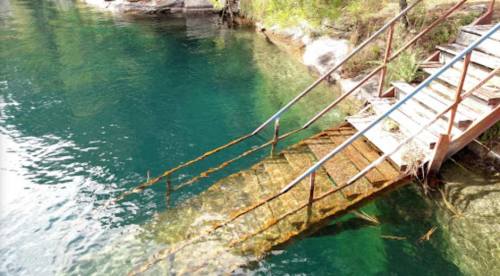 A rusted metal staircase leads into clear, turquoise water surrounded by rocks and greenery.