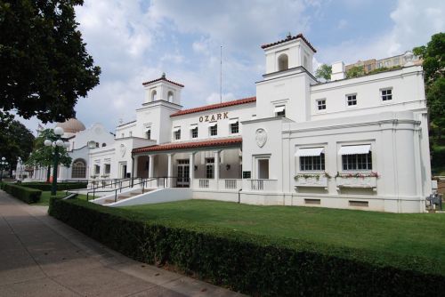 Historic white building with red-tiled roof, surrounded by green grass and trees, under a cloudy sky.