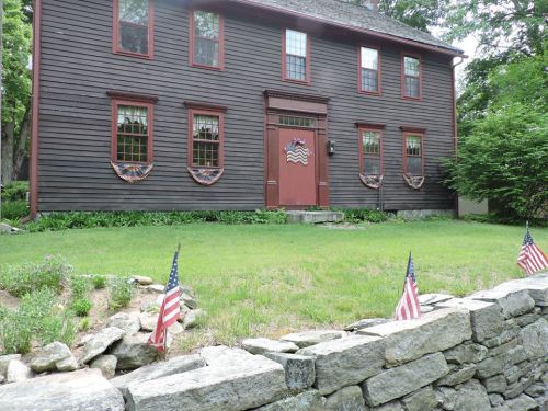 A brown wooden house with red accents, featuring decorative wreaths and small American flags in the front yard.