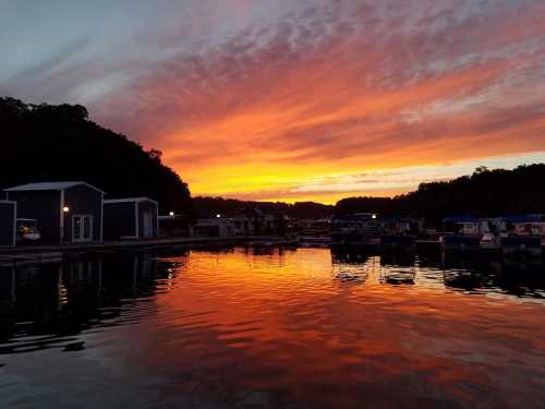 Sunset over a calm marina, with vibrant orange and purple skies reflecting on the water. Boathouses line the shore.