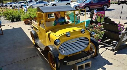 A child sits in a bright yellow vintage car outside, surrounded by flowers and other vehicles in a sunny setting.