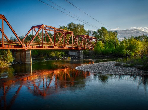 A red metal bridge spans a calm river, surrounded by lush greenery and a clear blue sky.