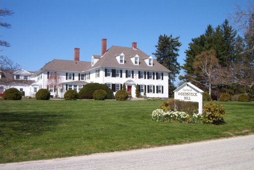 A large white colonial-style house with a manicured lawn and trees, featuring a sign for Woodstock Hill.