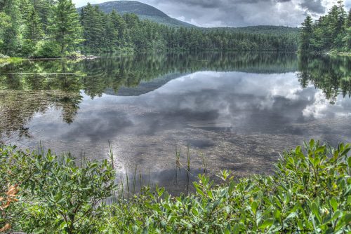 A serene lake surrounded by lush greenery and mountains, reflecting cloudy skies and trees in the calm water.