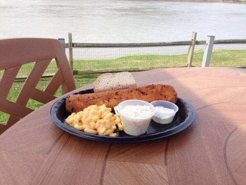 A plate with a breaded fish fillet, macaroni and cheese, coleslaw, and a slice of bread, set by a river.
