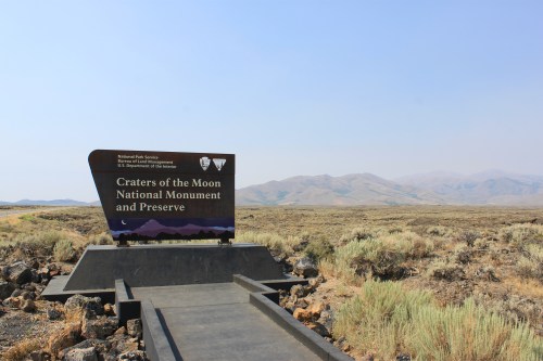 Sign for Craters of the Moon National Monument and Preserve, with a vast, arid landscape and mountains in the background.