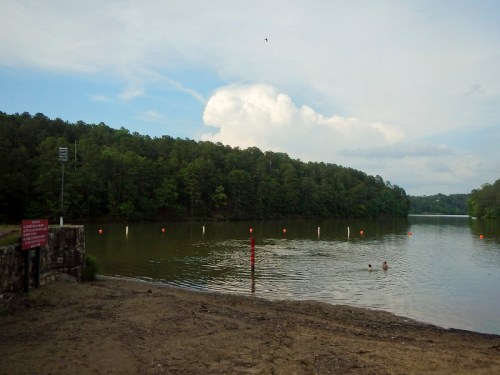 A serene lake surrounded by trees, with swimmers in the water and a cloudy sky above.