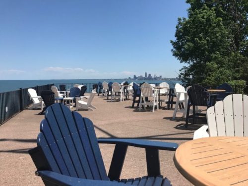 A lakeside patio with white and blue chairs, overlooking the water and a city skyline in the distance.