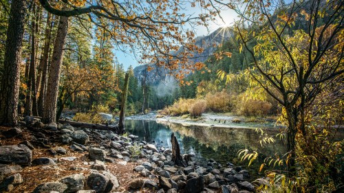 A serene river scene surrounded by autumn trees and rocky shores, with sunlight filtering through the foliage.