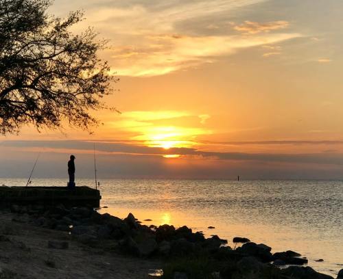 A person stands on a rocky shore at sunset, casting a fishing line into the calm water. Silhouetted against the vibrant sky.