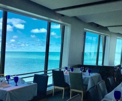 A seaside restaurant with large windows overlooking a blue ocean and sky, featuring tables set with blue glasses.